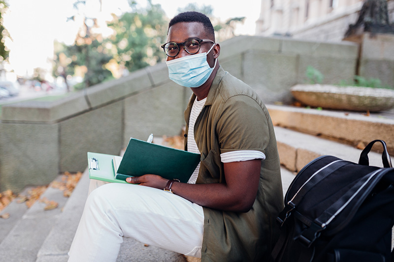 Male student sitting on a staircase in front of a university, taking notes in his note book and studying, while wearing a mask.