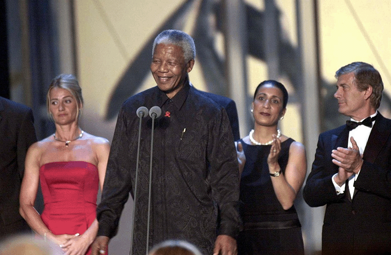 Nelson Mandela on stage giving a speech surrounded by Laureus Academy members Nadia Comaneci, Nawal El Moutawakel and Giacomo Agostini